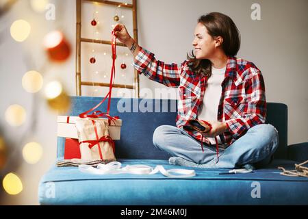 Junge Frau, die weihnachtsgeschenke packt und sich das rote Band ansieht. Stockfoto