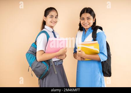 Fröhliche ländliche und moderne indische Schülerinnen in Schuluniform, die Bücher und Taschen halten und isoliert über beigefarbenen Hintergrund stehen Stockfoto