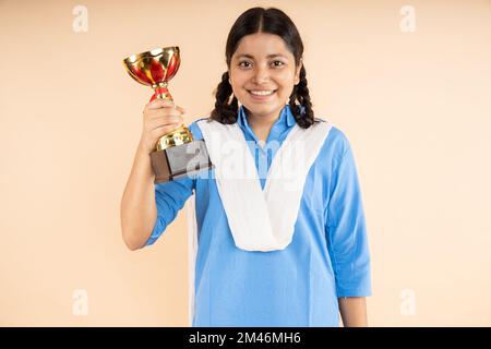 Glückliche, ländliche, indische Schülerin, die blaue Schuluniform trägt, hält die Siegestrophäe in der Hand, isoliert vor beigem Hintergrund, Sieger, Studio Stockfoto