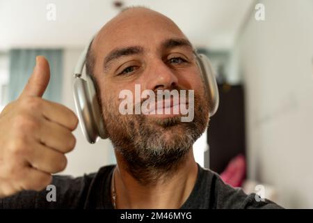Ein Mann mittleren Alters mit Bart sieht in die Kamera und zeigt Daumen-hoch-Gesten, während er zu Hause Musik über kabellose Kopfhörer hört Stockfoto