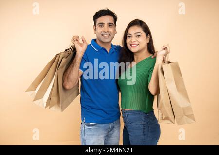 Glückliches, junges indisches Paar mit legeren Tüchern und braunen Papiertaschen, die isoliert auf beigefarbenem Studiohintergrund stehen. E-Commerce Stockfoto
