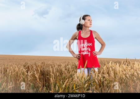 Frau, die auf einem Feld steht und Musik auf Kopfhörern hört, positive Vibes nur T-Shirt. Stockfoto