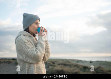 Eine Frau in Strickmode, die ein heißes Getränk genießt und dabei auf das Meer blickt. Stockfoto
