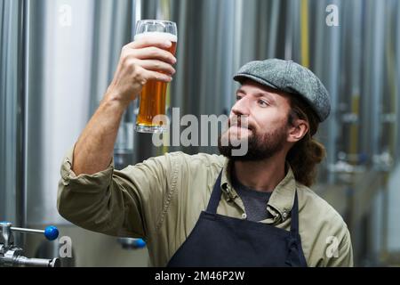 Glücklicher, bärtiger Brauereibesitzer, der sich ein Glas Bier ansah, das er gemacht hat Stockfoto