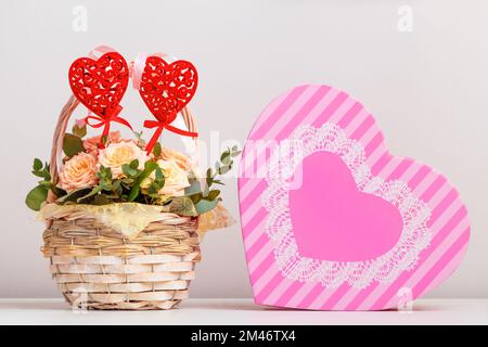 Zusammenstellung von Valentinstag, Muttertag, Geburtstag. Rosenblumen in einem Korb und eine rosa Schachtel mit einem herzförmigen Geschenk auf dem Tisch. Stockfoto