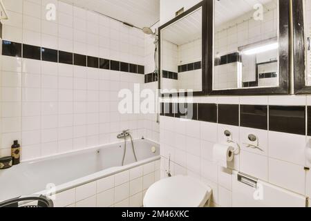 Badezimmer mit schwarz-weißen Fliesen an den Wänden, Badewanne, Toilette, Waschbecken, Spiegel und Duschkabine Stockfoto