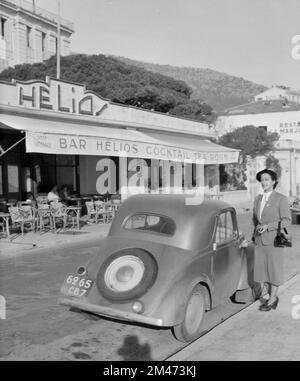 Junge Französin stellt den nächsten 5 2-türigen Saloon von Vintage Simca vor, der zwischen 1836 und 1948 in Nanterre Frankreich hergestellt wurde. Das französisch-italienische Auto war identisch mit dem Fiat 500 Topolina. Fotografiert vor dem Helios Restaurant am Meer oder am Ufer des Antibes Alpes-Maritimes Côte d'Azur oder der französischen Riviera France im Jahr 1951 Stockfoto