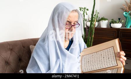 Alte muslimische Frau, die den Heiligen Koran zu Hause während des Ramadan-Monats las. Alte Frau trägt weißen Hijab und liest den Koran Stockfoto