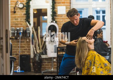 Friseurin während der Arbeit. Ein erfahrener Friseur schneidet die blonden Haare seines Klienten auf unkonventionelle Weise. Innenausstattung des Schönheitssalons. Hochwertiges Foto Stockfoto