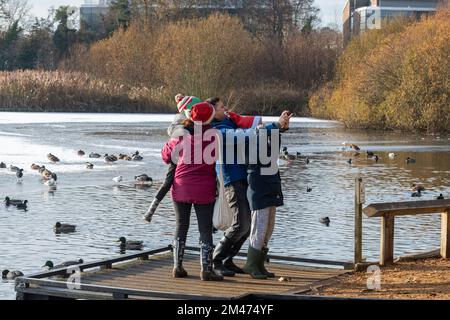 Familien genießen einen Winterspaziergang um einen gefrorenen Flottenteich, um Enten und Schwäne zu füttern und Selfies zu machen im Dezember 2022, Hampshire, England, Großbritannien Stockfoto