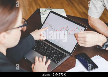 Mittelgroße Geschäftspartner arbeiten zusammen, während sie mit einer Kryptowährungstabelle am Laptop sitzen. Stockfoto