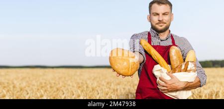 Ein Bauer mit einer Schürze steht auf einem Weizenfeld und hält Brotlaibe in der Hand. Stockfoto