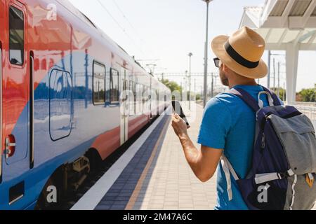 Mann mit Strohhut und Rucksack, der auf dem Bahnhofsplatz steht, mit Handy in der Hand und auf den Zug schaut. Stockfoto