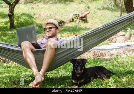 Ein Mann mit Hut, der auf einer Hängematte liegt und seinen Laptop benutzt, ein Hund, der neben dem Besitzer auf dem Boden liegt. Stockfoto