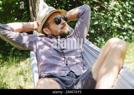 Bärtiger Mann mit Hut und Sonnenbrille, der auf einer Hängematte im grünen Garten liegt. Stockfoto