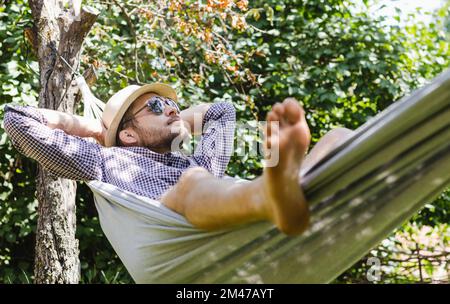 Bärtiger Mann mit Hut und Sonnenbrille, der in der Hängematte im grünen Garten liegt. Stockfoto