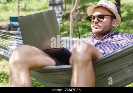 Ein Mann mit Hut, der auf einer Hängematte liegt und auf seinen Laptop schaut. Stockfoto