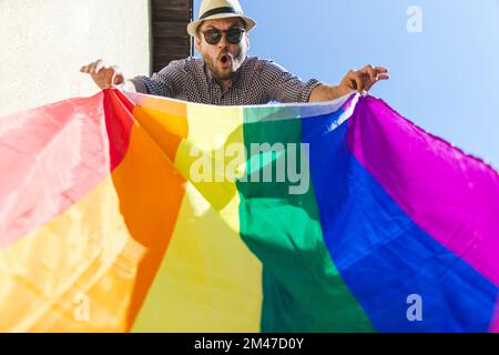 Ein positiver Mann mit Hut, der eine große LGBT-Regenbogenfahne in den Händen hält. Stockfoto