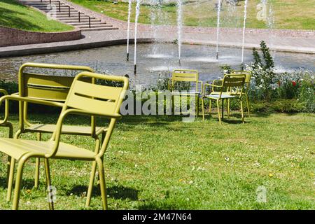 Bewegliche Stühle im öffentlichen Raum einer modernen Stadt. Kostenlose grüne Stahlstühle im Park auf dem Rasen neben dem Teich. Das Konzept moderner Stadtlandschaften Stockfoto