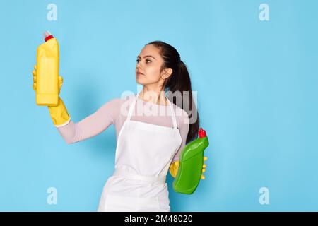 Frau in Handschuhen und Reinigungsschürze wählt zwischen zwei Reinigungsprodukten Stockfoto