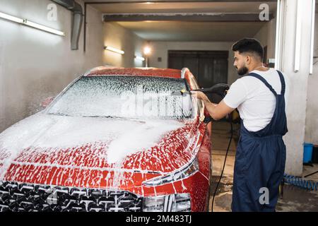 Porträt eines jungen bärtigen Mannes in Arbeitskleidung, Autowaschanlagenangestellten, Waschen der Windschutzscheibe unter einer Hochdruck-Seifenwasserpistole bei einem professionellen Autowaschanlagenservice. Stockfoto