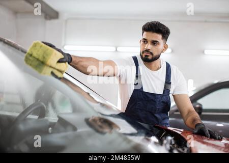 Junger bärtiger Mann, Autowaschanlage, trägt T-Shirt und Overalls und reinigt die Windschutzscheibe des Autos mit Hilfe von speziellem Schaum und Schwamm. Das Auto von Hand waschen, Schwamm und Schaumstoffblasen. Stockfoto