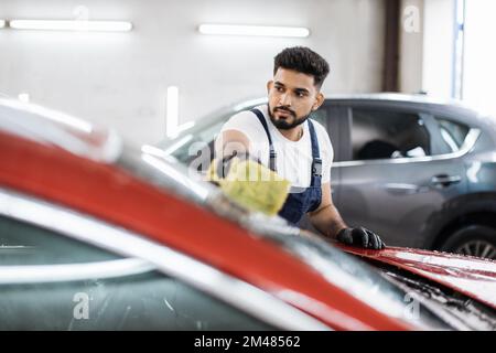 Junger bärtiger Mann, Autowaschanlage, trägt T-Shirt und Overalls und reinigt die Windschutzscheibe des Autos mit Hilfe von speziellem Schaum und Schwamm. Das Auto von Hand waschen, Schwamm und Schaumstoffblasen. Stockfoto