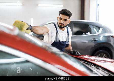 Junger bärtiger Mann, Autowaschanlage, trägt T-Shirt und Overalls und reinigt die Windschutzscheibe des Autos mit Hilfe von speziellem Schaum und Schwamm. Das Auto von Hand waschen, Schwamm und Schaumstoffblasen. Stockfoto