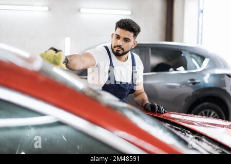 Junger bärtiger Mann, Autowaschanlage, trägt T-Shirt und Overalls und reinigt die Windschutzscheibe des Autos mit Hilfe von speziellem Schaum und Schwamm. Das Auto von Hand waschen, Schwamm und Schaumstoffblasen. Stockfoto