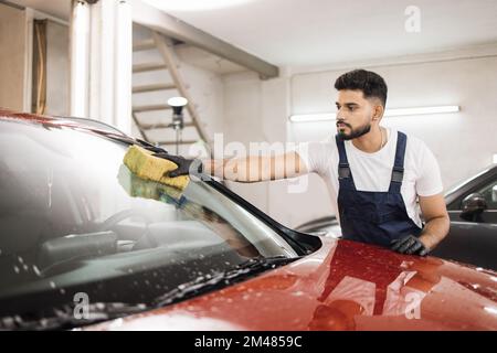 Selbstbewusster junger Mann, Autowaschanlage, trägt T-Shirt und Overalls und reinigt die Windschutzscheibe des Autos mit Hilfe von speziellem Schaum und Schwamm. Das Auto von Hand waschen, Schwamm und Schaumstoffblasen. Stockfoto
