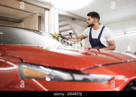 Selbstbewusster junger Mann, Autowaschanlage, trägt T-Shirt und Overalls und reinigt die Windschutzscheibe des Autos mit Hilfe von speziellem Schaum und Schwamm. Das Auto von Hand waschen, Schwamm und Schaumstoffblasen. Stockfoto