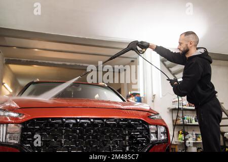 Das Fahrzeug mit Hochdruckwasser reinigen. Attraktiver junger Mann, der ein modernes rotes Auto unter Hochdruck wäscht. Stockfoto