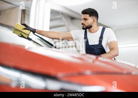 Selbstbewusster junger Mann, Autowaschanlage, trägt T-Shirt und Overalls und reinigt die Windschutzscheibe des Autos mit Hilfe von speziellem Schaum und Schwamm. Das Auto von Hand waschen, Schwamm und Schaumstoffblasen. Stockfoto