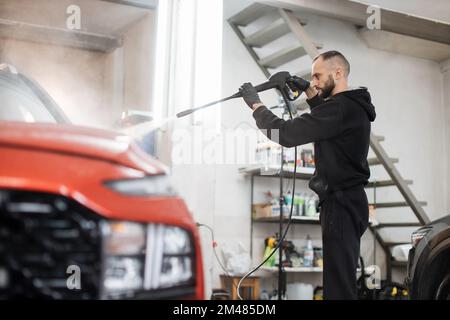 Das Fahrzeug mit Hochdruckwasser reinigen. Attraktiver junger Mann, der die moderne rote Motorhaube unter Hochdruck wäscht. Stockfoto