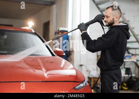 Das Fahrzeug mit Hochdruckwasser reinigen. Attraktiver junger Mann, der die moderne rote Motorhaube unter Hochdruck wäscht. Stockfoto