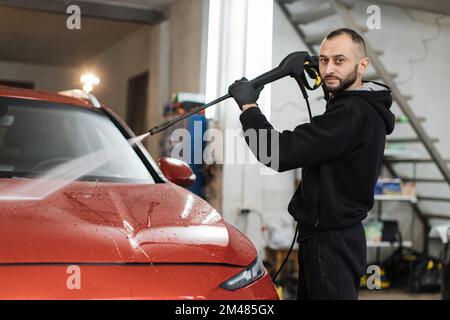 Das Fahrzeug mit Hochdruckwasser reinigen. Attraktiver junger Mann, der die moderne rote Motorhaube unter Hochdruck wäscht. Stockfoto