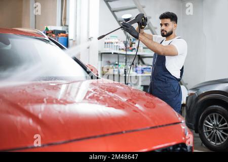 Das Fahrzeug mit Hochdruckwasser reinigen. Attraktiver junger Mann, der die moderne rote Motorhaube unter Hochdruck wäscht. Stockfoto