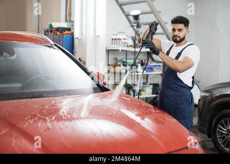 Das Fahrzeug mit Hochdruckwasser reinigen. Attraktiver junger Mann, der die moderne rote Motorhaube unter Hochdruck wäscht. Stockfoto