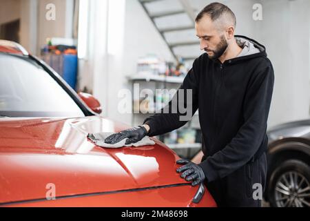 Fahrzeugdetails und Reinigungskonzept. Gut aussehender junger Autowaschanlagenarbeiter, der spezielle Kleidung und Handschuhe trägt, die Motorhaube mit Mikrofasertuch reinigt und poliert. Stockfoto