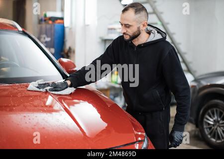 Fahrzeugdetails und Reinigungskonzept. Gut aussehender junger Autowaschanlagenarbeiter, der spezielle Kleidung und Handschuhe trägt, die Motorhaube mit Mikrofasertuch reinigt und poliert. Stockfoto