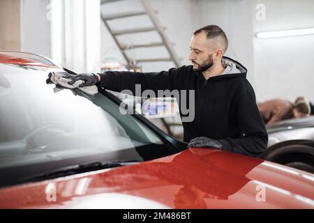 Fahrzeugdetails und Reinigungskonzept. Gut aussehender junger Autowaschanlagenarbeiter, trägt schwarze Handschuhe, reinigt und poliert Autofenster mit Mikrofasertuch. Stockfoto