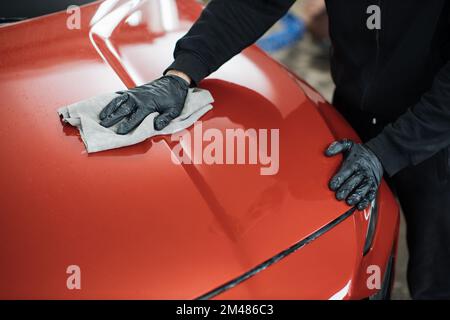 Nahaufnahme der Hände eines männlichen Autowaschanlagenarbeiters, der spezielle Kleidung und Handschuhe trägt, wischt und poliert die Motorhaube mit einem Mikrofasertuch. Fahrzeugdetails und Reinigungskonzept. Stockfoto