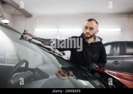 Fahrzeugdetails und Reinigungskonzept. Gut aussehender junger Autowaschanlagenarbeiter, trägt schwarze Handschuhe, reinigt und poliert Autofenster mit Mikrofasertuch. Stockfoto