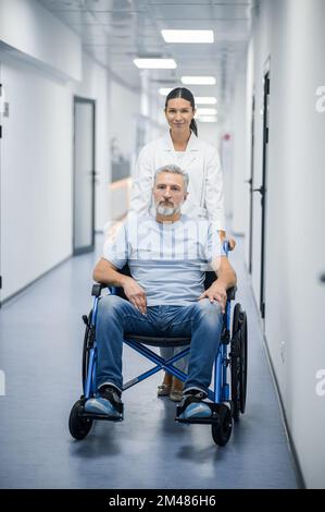 Dunkelhaarige Krankenschwester, die den Rollstuhl mit einem Patienten trägt Stockfoto