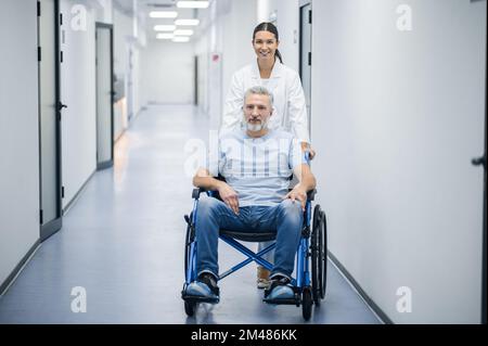 Dunkelhaarige Krankenschwester, die den Rollstuhl mit einem Patienten trägt Stockfoto