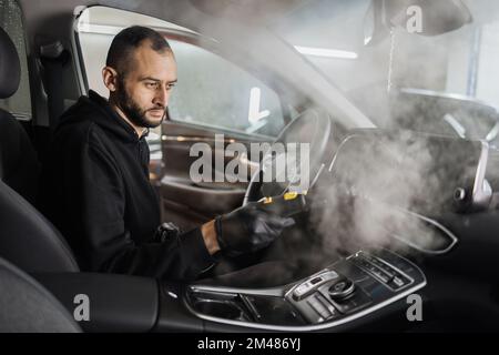 Automatischer Reinigungsservice und Detaillierungskonzept. Ein gutaussehender bärtiger Mann, der den Innenraum des Autos mit heißem Dampfreiniger reinigt. Selektiver Fokus auf Männergesicht. Stockfoto