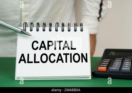 Geschäftskonzept. Auf einer grünen Oberfläche befindet sich ein Taschenrechner, ein Mann zeigt mit einem Stift auf ein Notizbuch mit der Aufschrift "Kapitalzuweisung" Stockfoto
