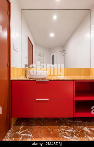 Weißes Spüle über Kopf in einem roten Schrank in einem hellen weißen und gelben Badezimmer Weiße und gelbe Fliesen an der Wand und brauner Boden aus Porzellan-Steingut Stockfoto