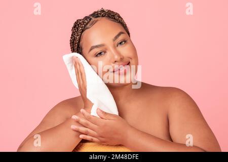 Hygienekonzept. Wunderschöne schwarze Frau in Übergröße trocknet sauberes Gesicht mit einem Handtuch, steht über pinkfarbenem Studiohintergrund Stockfoto