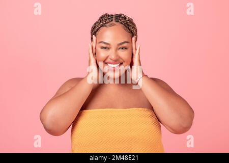 Porträt einer aufgeregten, schwarzen, körperpositiven Frau, die ihr Gesicht berührt und ihre glatte Haut genießt, rosa Studiohintergrund Stockfoto